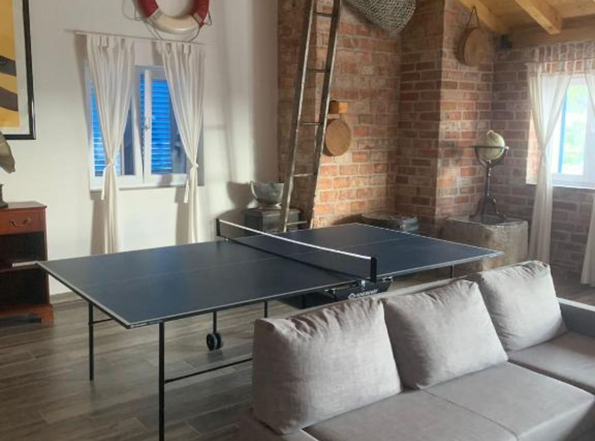 A ping pong table in a room with brick walls, a sofa, and a window with curtains.
