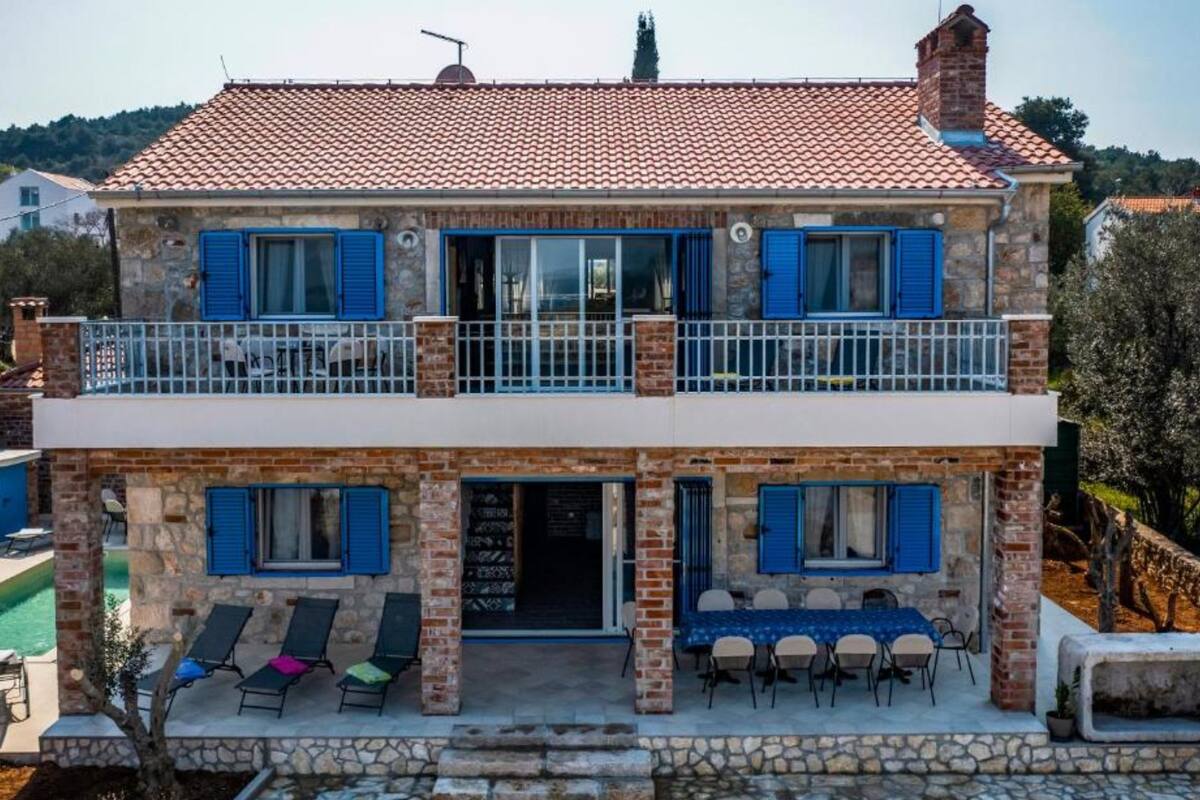 A two-story stone house with blue shutters, a balcony, and a pool.