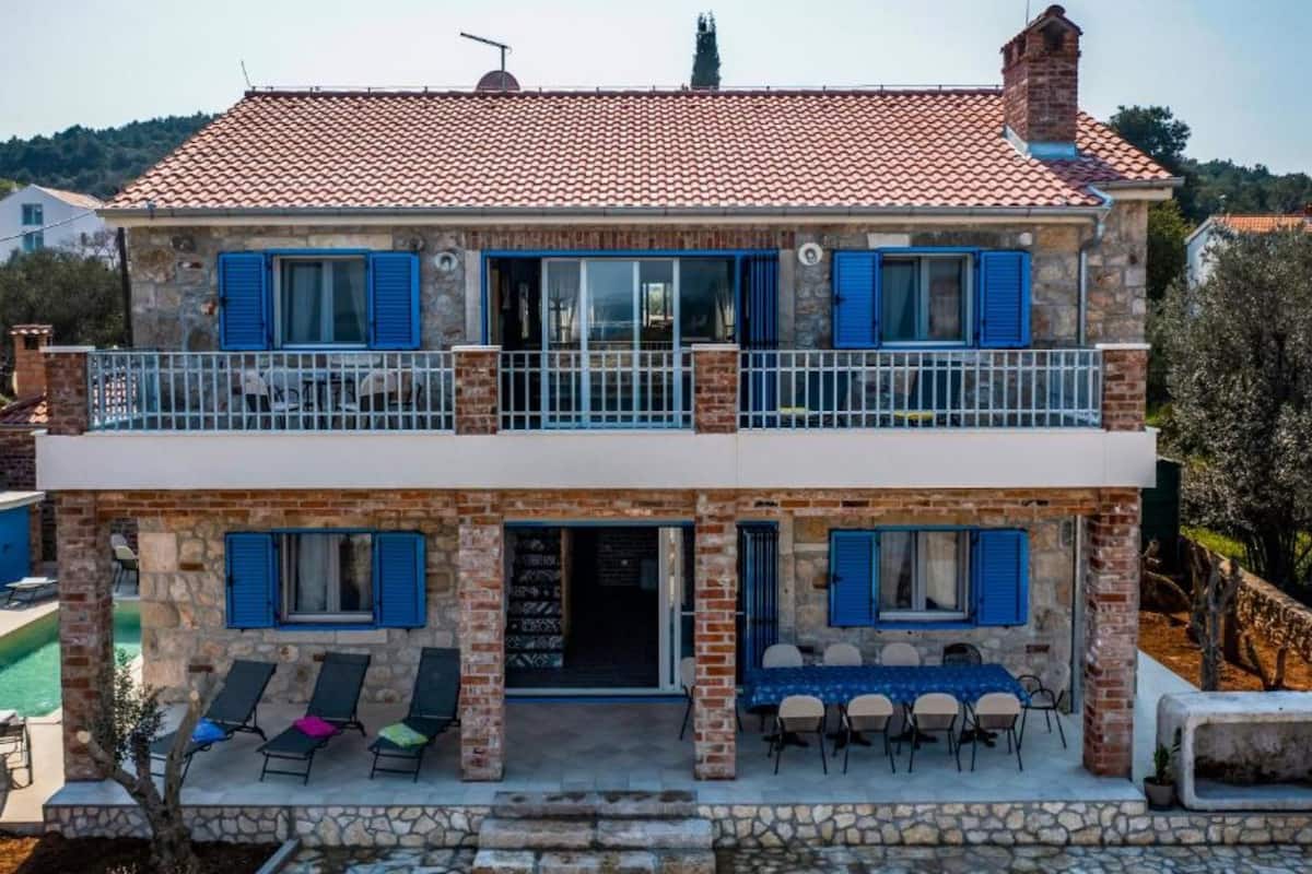 A two-story stone house with blue shutters, a balcony, and a pool.