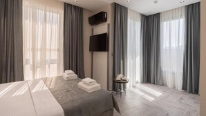 A hotel room with a bed, a TV mounted on the wall, a small table with a cup and saucer, and large windows with curtains. - Hotel D'iman (Bar)