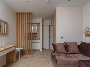 Una habitación moderna con separador de madera, un escritorio con silla, un sofá con cojines y una cocina al fondo.