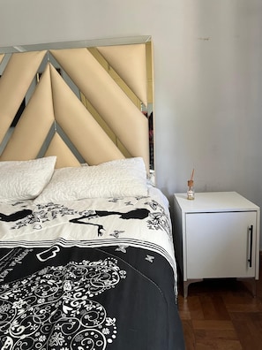 Sängyssä on kuviollinen päiväpeite, geometrisesti kuvioitu pääty ja yöpöydällä on pullo.