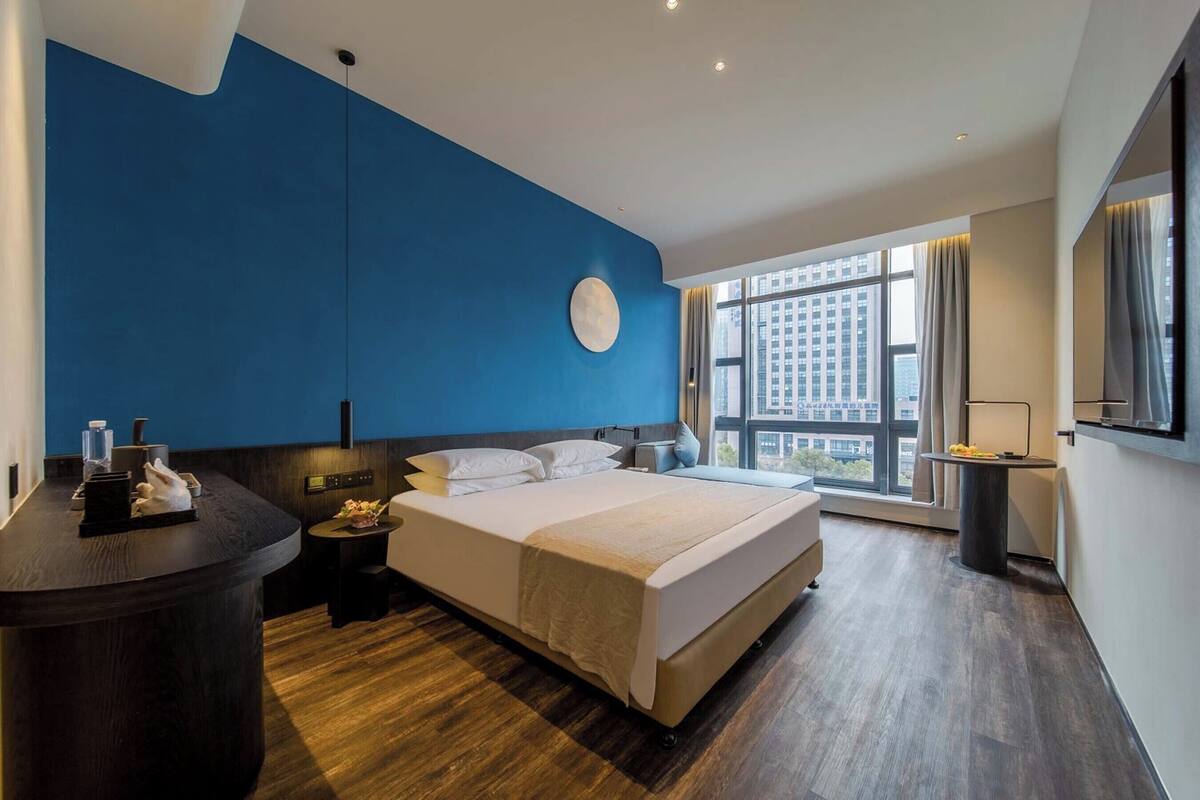 A modern hotel room with a large bed, a desk, a round wall clock, and a view of the city through large windows.