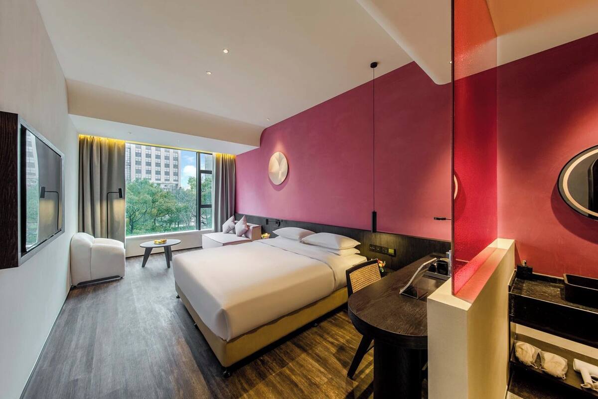 A modern hotel room with a large bed, a desk, a chair, a round table, a TV, and a large window with curtains.