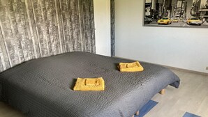 A bed with a textured grey bedspread, two folded towels, a blue rug, and a city street view poster on the wall. - Large family home in Normandy (Ganzeville)