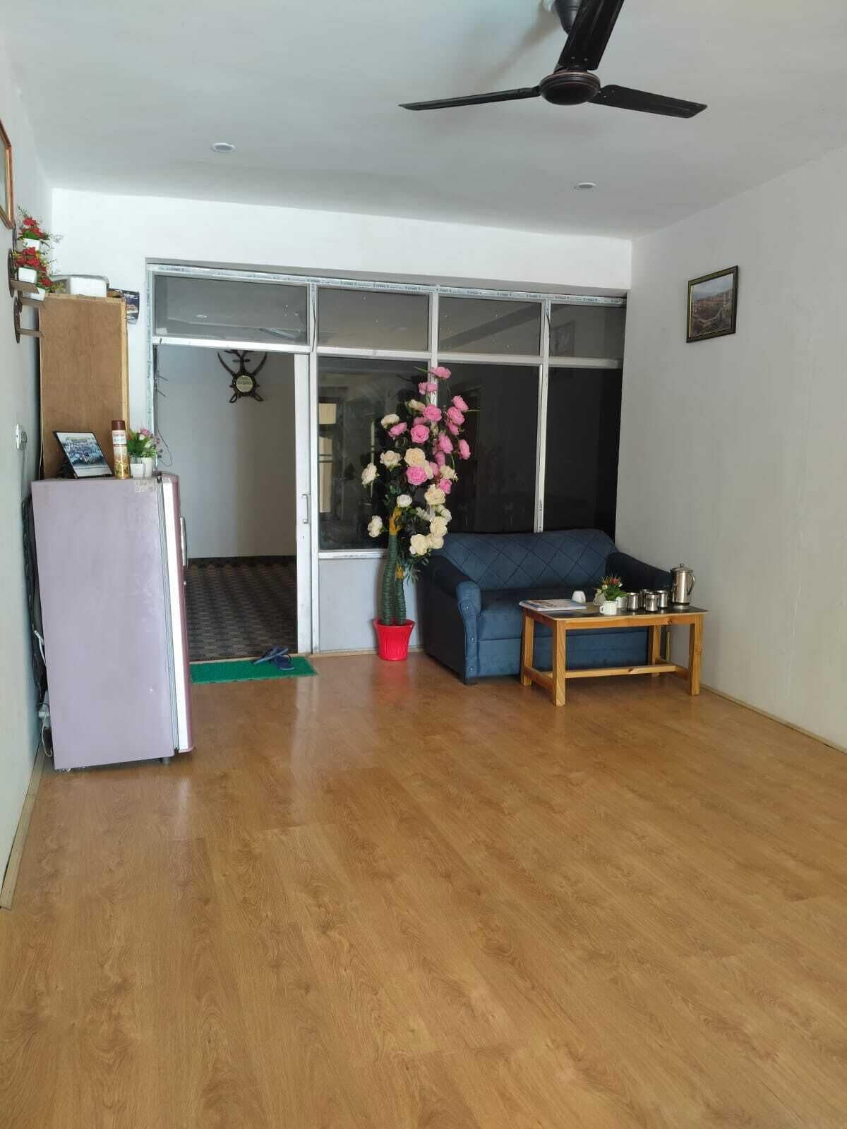 Un salón con suelo de madera, un sofá azul, una mesita con objetos, un refrigerador, una planta en maceta y una ventana con vistas.