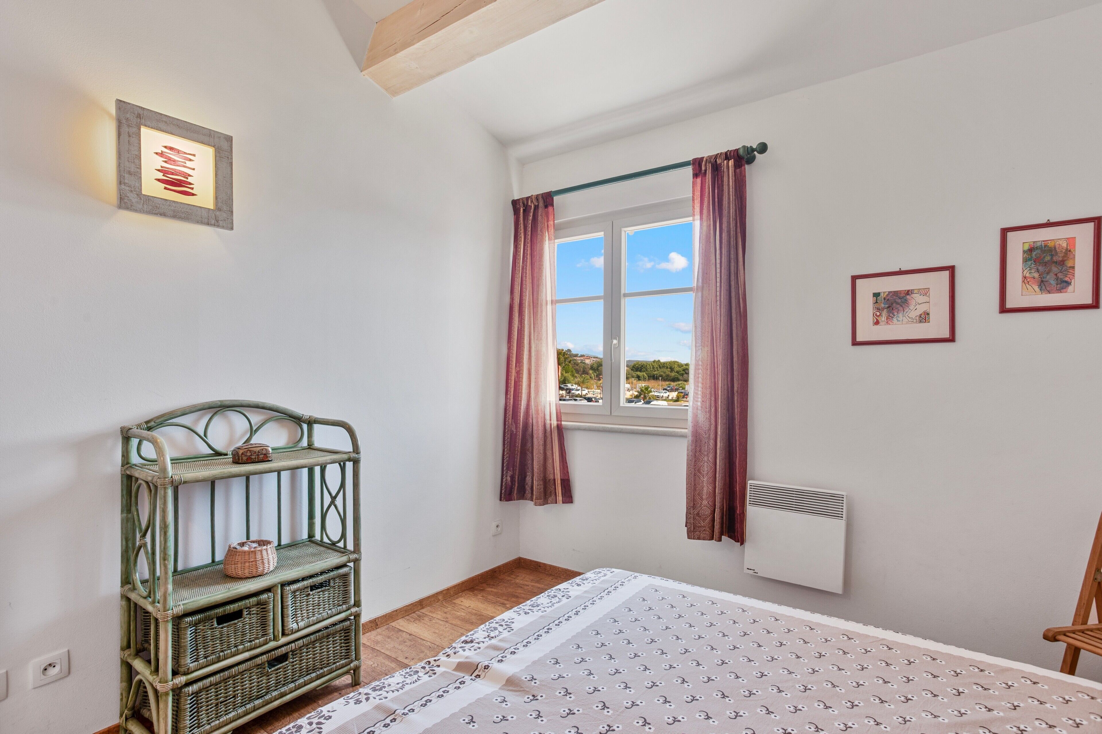 Une chambre à coucher comprenant un lit, une fenêtre avec des rideaux, un climatiseur fixé au mur et un meuble en osier.