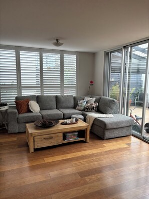 A modern living room with a sectional sofa, a wooden coffee table, and large windows with blinds. - Edithvale Beach House (Edithvale)