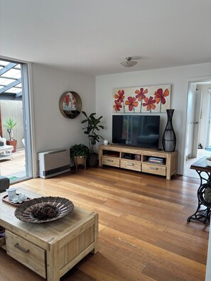 A living room with a wooden TV stand, a flat-screen TV, a large floral painting, a mirror, and a potted plant. - Edithvale Beach House (Edithvale)