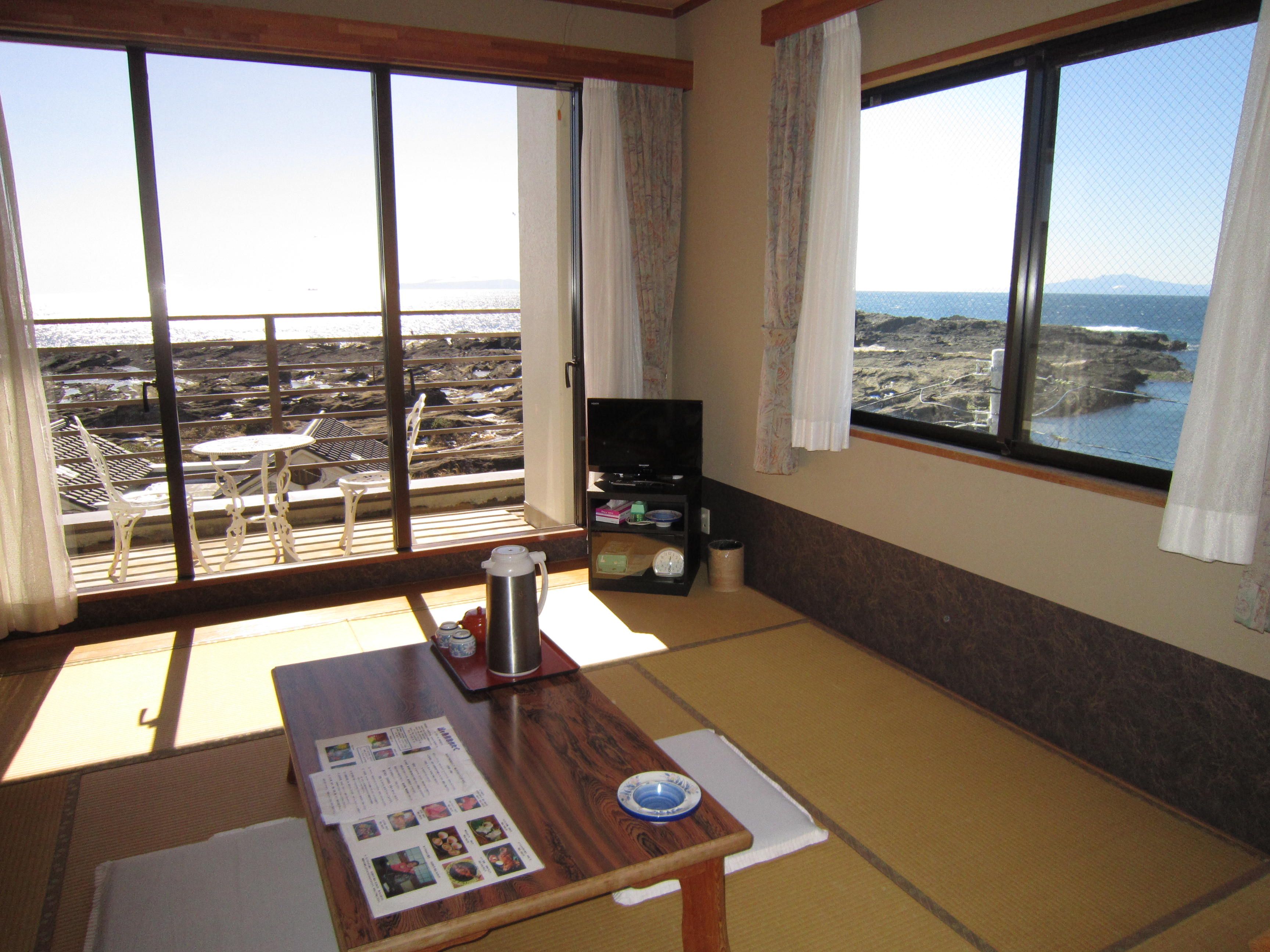 A room with tatami flooring, a low table with a tray, a television, and a view of a rocky coastline through large windows.