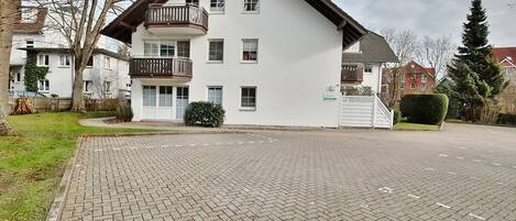 Un edificio residencial de varios pisos con balcón, entrada de ladrillo y vegetación circundante.