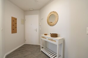 Ein Flur mit einer weißen Tür, einer Holztruhe mit Haken, einem gewebten Spiegel und einem Konsolentisch mit Körben.