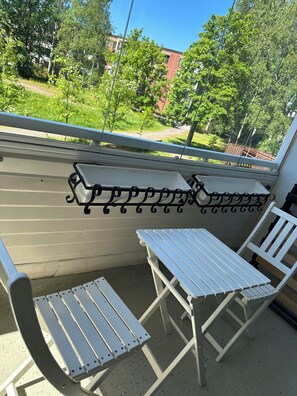 A balcony with two white metal chairs and a small table, overlooking a green area with trees and a building. - NOT LISTED (Helsinki)