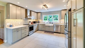 A modern kitchen with light grey cabinets, stainless steel appliances, and a large window providing a view of greenery. - 3 Acres, Mtn Views, Hot Tub, Steps to Winery (Richmond)