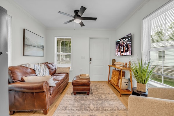 A living room with a brown leather couch, a ceiling fan, a television, and a large window with blinds.