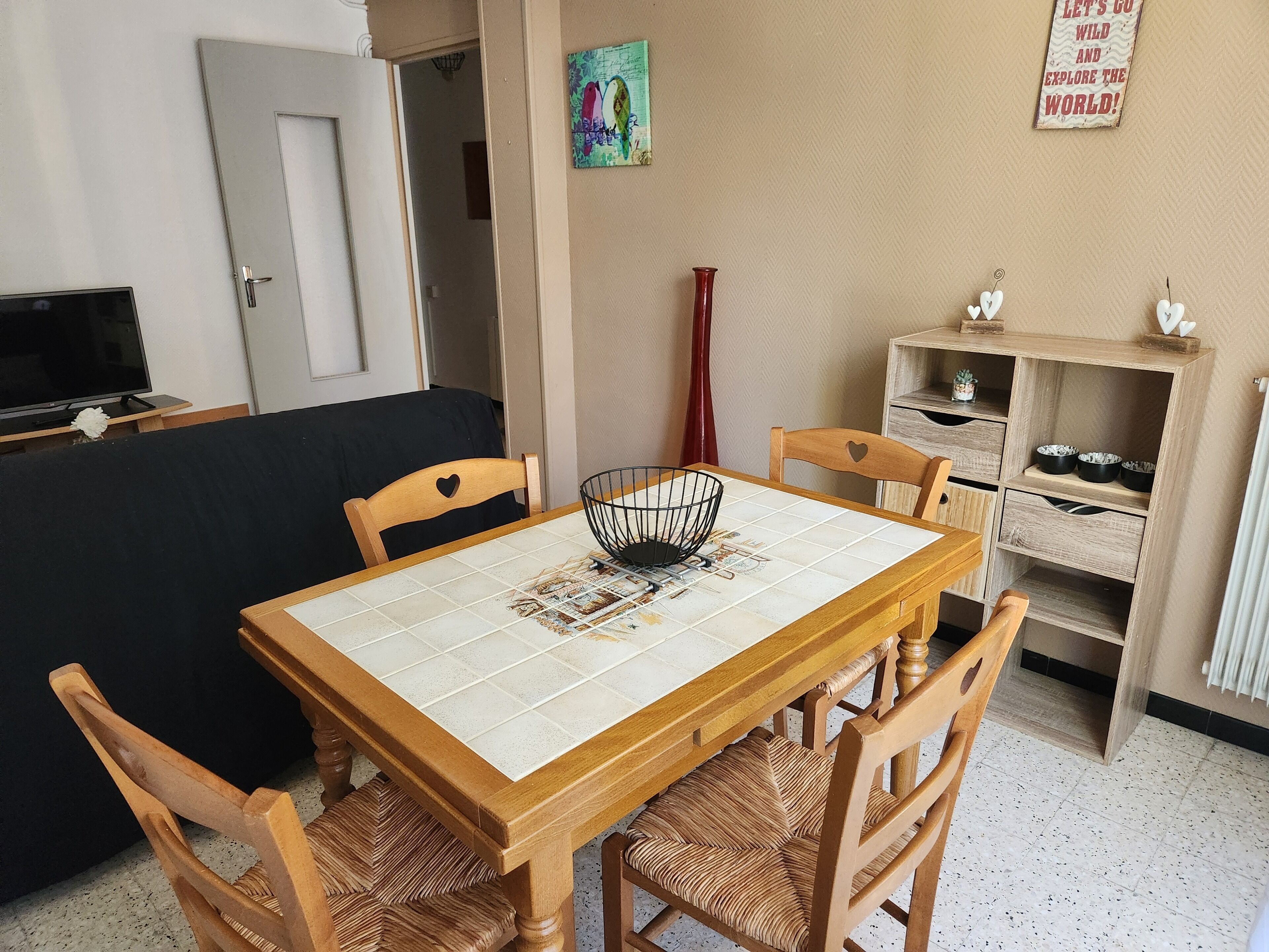 A dining area with a wooden table, four chairs, a TV, a painting, and a shelf with bowls.