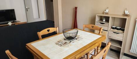 A dining area with a wooden table, four chairs, a TV, a painting, and a shelf with bowls.