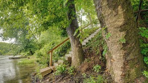 Una scala di legno conduce a una piattaforma in pietra vicino a un fiume, circondata da una vegetazione lussureggiante.