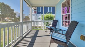 A covered porch with two wicker chairs and a table, a white railing, and a view of a blue house with a white fence. - Steps to Ocean! Family-Friendly Carolina Beach Gem (Carolina Beach)