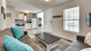 A living room with a sofa, a coffee table, a ceiling fan, a kitchen area with appliances, and a large window. - Steps to Ocean! Family-Friendly Carolina Beach Gem (Carolina Beach)