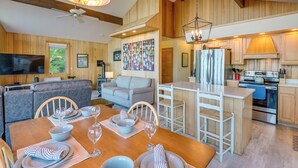 Une cuisine moderne avec des meubles en bois, un coin repas avec une table en bois dressée pour quatre personnes, et un salon avec un canapé et une télévision.