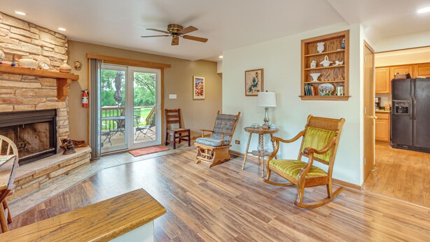 A spacious living area with a stone fireplace, wooden flooring, a ceiling fan, and a view through glass doors to an outdoor patio. - Fall Escape! Waterfront Chain O'Lakes Hideaway (Spring Grove)
