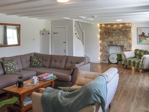 Ein Wohnzimmer mit einem braunen Ecksofa, einem weißen Sessel, einem Couchtisch mit Büchern und einer Kaffeekanne, einem Kamin mit Lichterketten und einer Treppe.