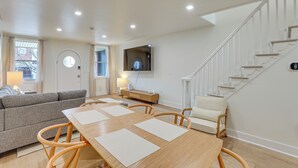 Un salon moderne avec une table à manger en bois, un canapé gris, un fauteuil blanc, un téléviseur fixé au mur et un escalier.