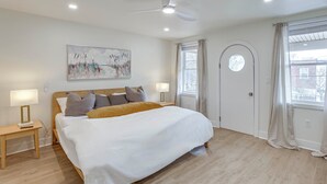 Une chambre à coucher comprenant un lit, des tables de chevet, un ventilateur de plafond, une fenêtre avec des rideaux et une porte.