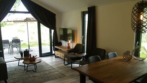 Uma sala de estar moderna com uma mesa de jantar de madeira, assentos confortáveis, uma TV de tela plana e uma grande porta de vidro deslizante que dá para uma área externa.