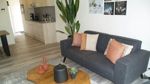 Uma sala de estar moderna com um sofá cinza, uma mesa de centro de madeira, uma planta em vaso e uma cozinha ao fundo.