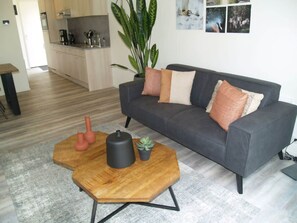 Ein modernes Wohnzimmer mit einem grauen Sofa, einem Holztisch, einer Topfpflanze und einer Küche im Hintergrund.