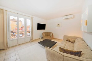 En stue med sofa, sofabord, stol, fjernsyn og en balkon med udsigt til bygninger.