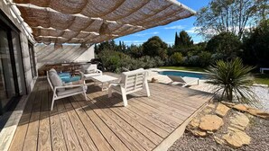 En træterrasse med udendørs møbler, en pool og et pergola der giver skygge.