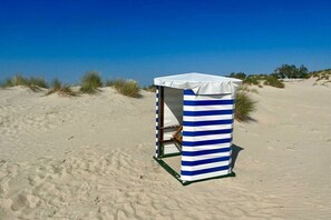 Eine Strandhütte mit gestreiftem Markisenvorzelt, gelegen an einem Sandstrand mit Dünen im Hintergrund.