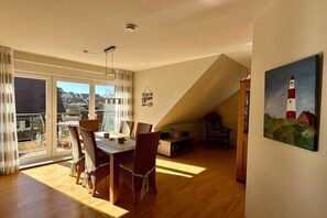 A dining area with a table and chairs, a painting of a lighthouse, and a view through large windows. - Holiday apartment with 2 bedrooms (Borkum)
