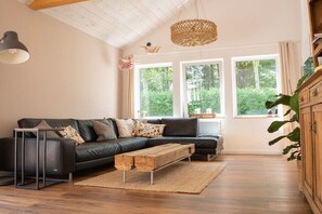 Ein modernes Wohnzimmer mit einem schwarzen Ledersofa, einem Holztisch, einer gewobenen Pendelleuchte und großen Fenstern mit Blick auf Grünflächen.