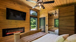 Een slaapkamer in een houten hutje, met een open haard, een bed, een plafondventilator en een aan de muur bevestigde televisie.