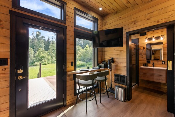 Interior de una cabaña de madera con comedor, cocina y un amplio ventanal con vistas al bosque.