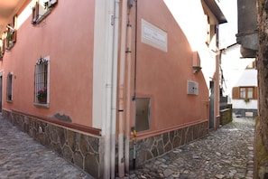 Ein Gebäude mit steinernem Sockel und einer rosafarbenen Außenwand, eine Schrifttafel mit der Aufschrift 'Via Dotti Baggiolini' und eine enge, gepflasterte Straße.