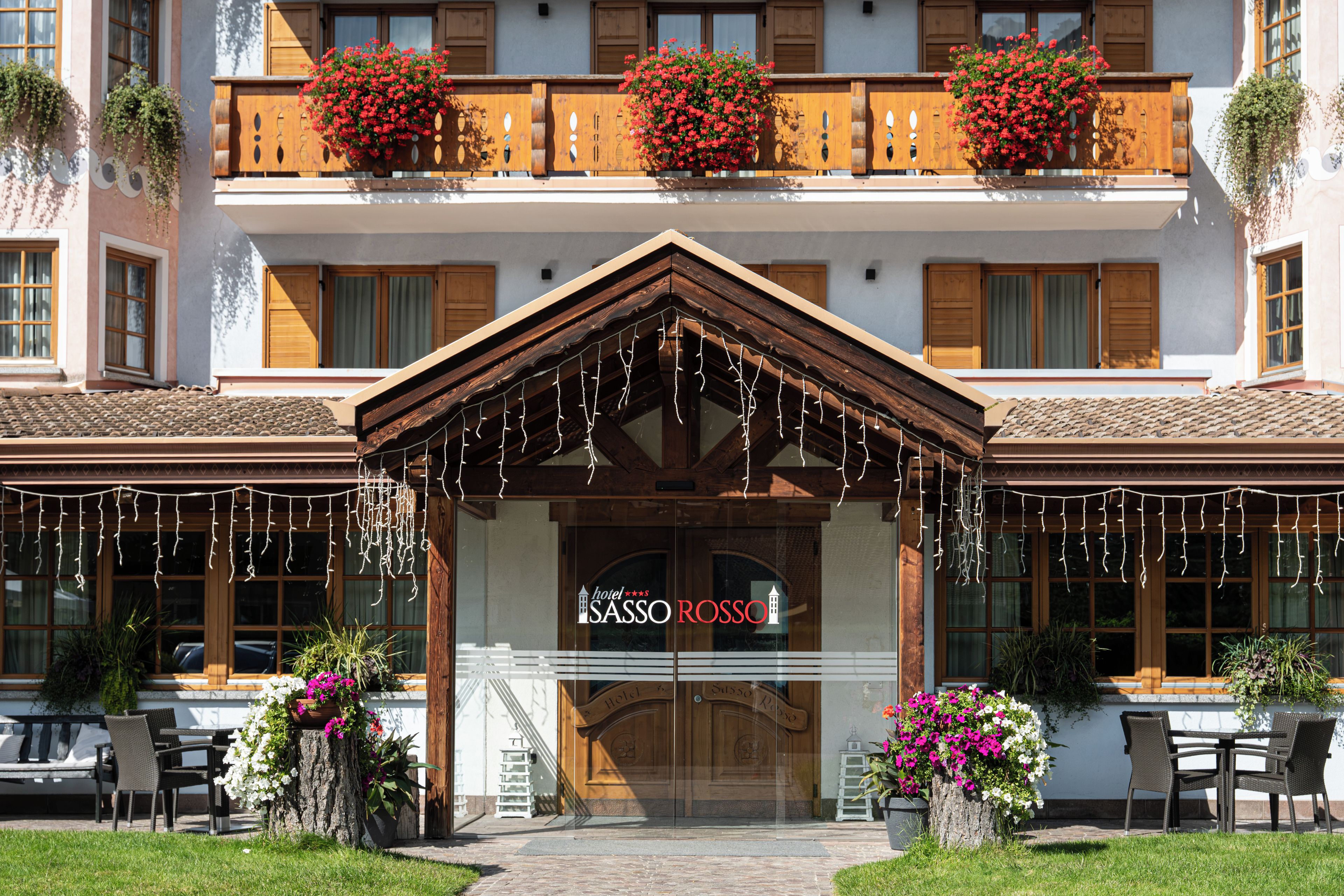 Entrada de hotel con fachada de madera, un letrero que dice 'Sasso Rosso' y mobiliario exterior con plantas en macetas.