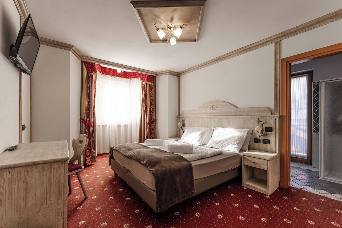 Una habitación de hotel con cama, mesita de noche, televisor y una alfombra roja estampada.