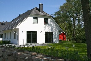 Ein modernes, zweistöckiges Haus mit geneigtem Dach, großen Fenstern und einem roten Bauernhaus im Hintergrund.