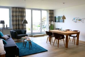 Ein modernes Wohnzimmer mit einer Couch, einem Esstisch mit Stühlen, einem an der Wand befestigten Fernseher und großen Fenstern mit Vorhängen.
