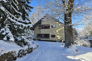 Um edifício de dois andares com paisagem coberta de neve, entrada visível e uma árvore com um comedouro para pássaros.