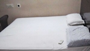 Una cama individual con colchón y almohadas blancos, una mesita de noche pequeña y un enchufe de pared.