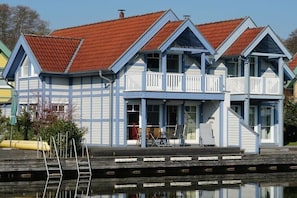 Ein Gebäude am Wasser mit rotem Ziegeldach, weiß-blauer Fassade und einem Balkon mit Stühlen.