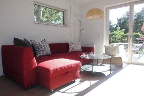 Ein rotes Sofa, ein Glastisch und ein Korbstuhl in einem Raum mit einem großen Fenster und einer Stehlampe.