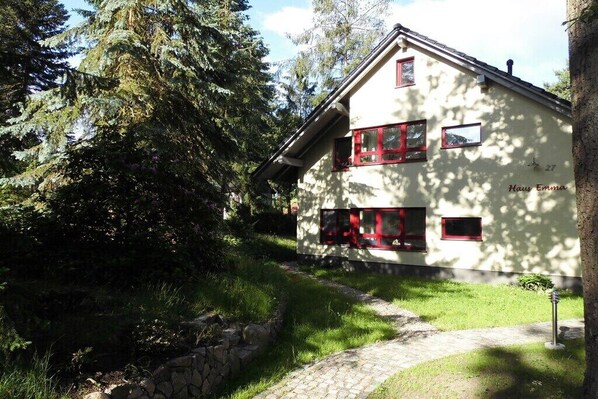 Une maison à deux étages, avec un chemin de pierre menant à l’entrée, entourée de verdure et d’arbres.