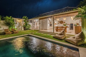 A pool area with lounge chairs, a dining area with a table and chairs, and a kitchen visible through an open door. - 1BR Romantic Getaway in Ubud, Villa Lojo with Private Pool (Gianyar)